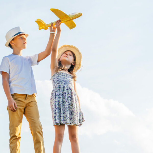 Portrait of children boy and girl playing outdoors. Light sunny day. Cheerful and calm concept. Fresh air in countryside. Kids wearing hats and playing flying yellow toy airplane. Planning travelling