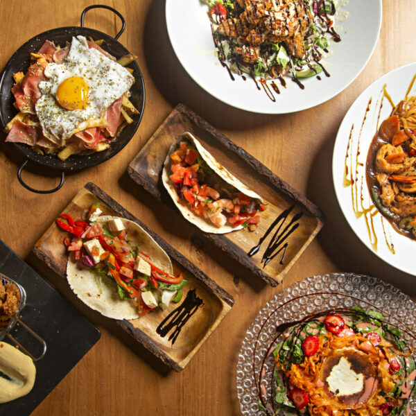 Varied food dishes of the world on wooden table, eggs, salad, chicken, tacos.